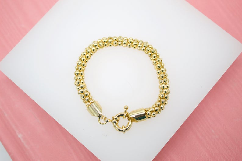 A gold beaded popcorn bracelet rests on a white display surface against a pink background.