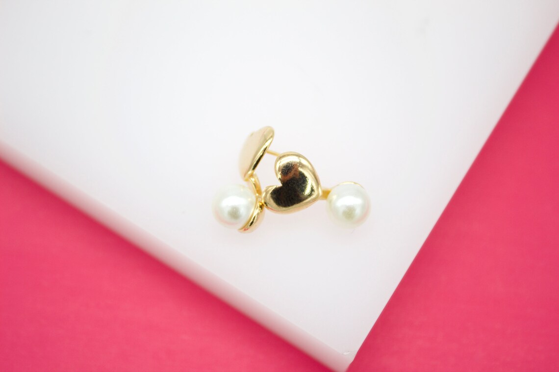 A pair of delicate gold heart-shaped earrings features dangling white pearls against a white and pink background.