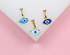 Three gold evil eye charms in light blue, dark blue, and white display detailed enamel work against a pink and white background.