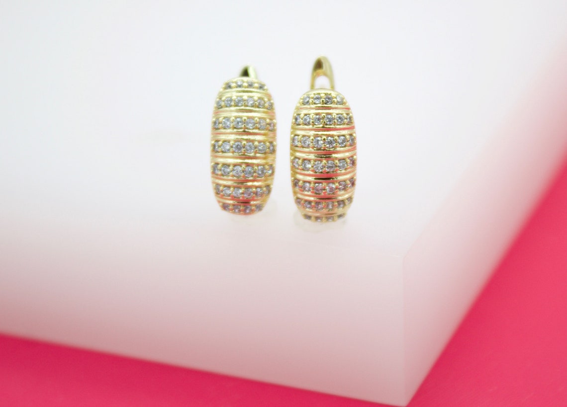 A pair of gold huggie hoop earrings featuring rows of sparkling cubic zirconia stones rests upright on a white display surface against a pink background.