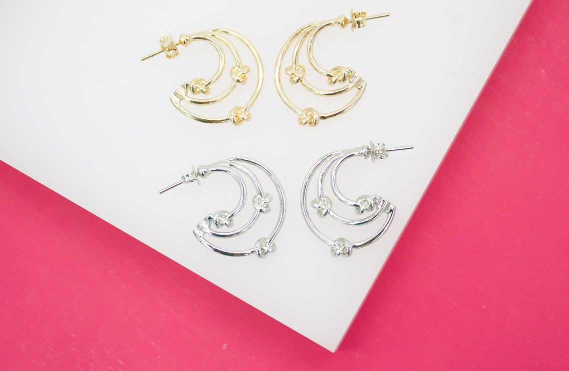 Two pairs of multi-strand hoop earrings with knot details rest on a white surface, featuring gold earrings on top and silver earrings below.