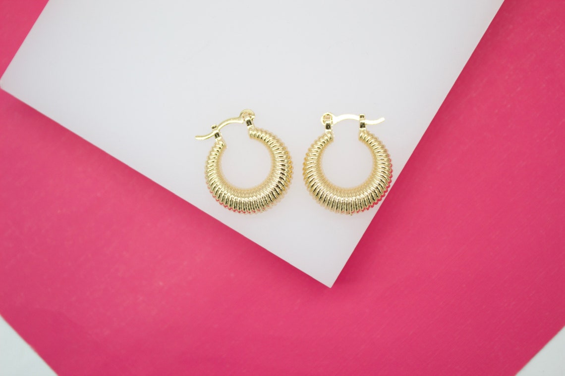 A pair of thick gold hoop earrings with a ribbed texture rest on a white surface against a bright pink background.