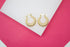 A pair of thick gold hoop earrings with a ribbed texture rest on a white surface against a bright pink background.