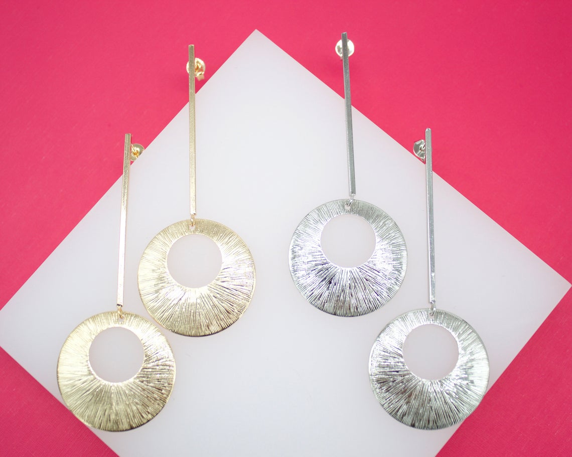 Two pairs of long, dangling geometric earrings featuring textured gold and silver circular pendants display on a white display against a vibrant pink background.