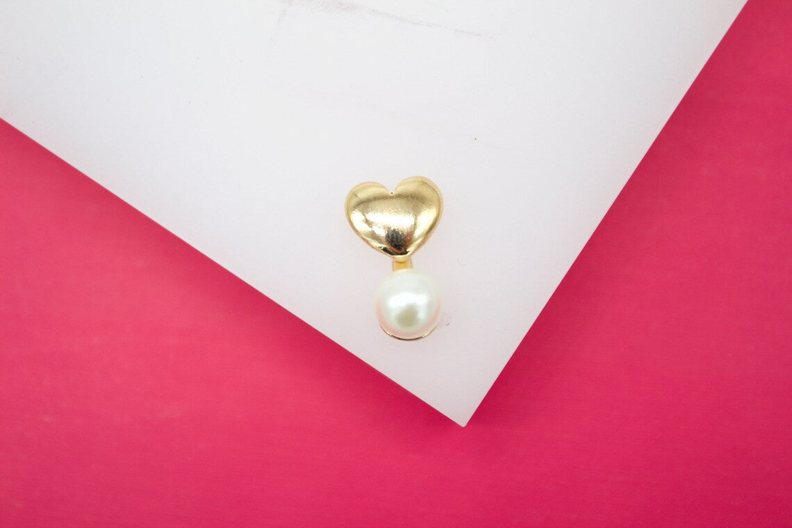 A chic gold heart-shaped stud earring with a dangling white pearl rests on a white display surface against a vibrant pink background.