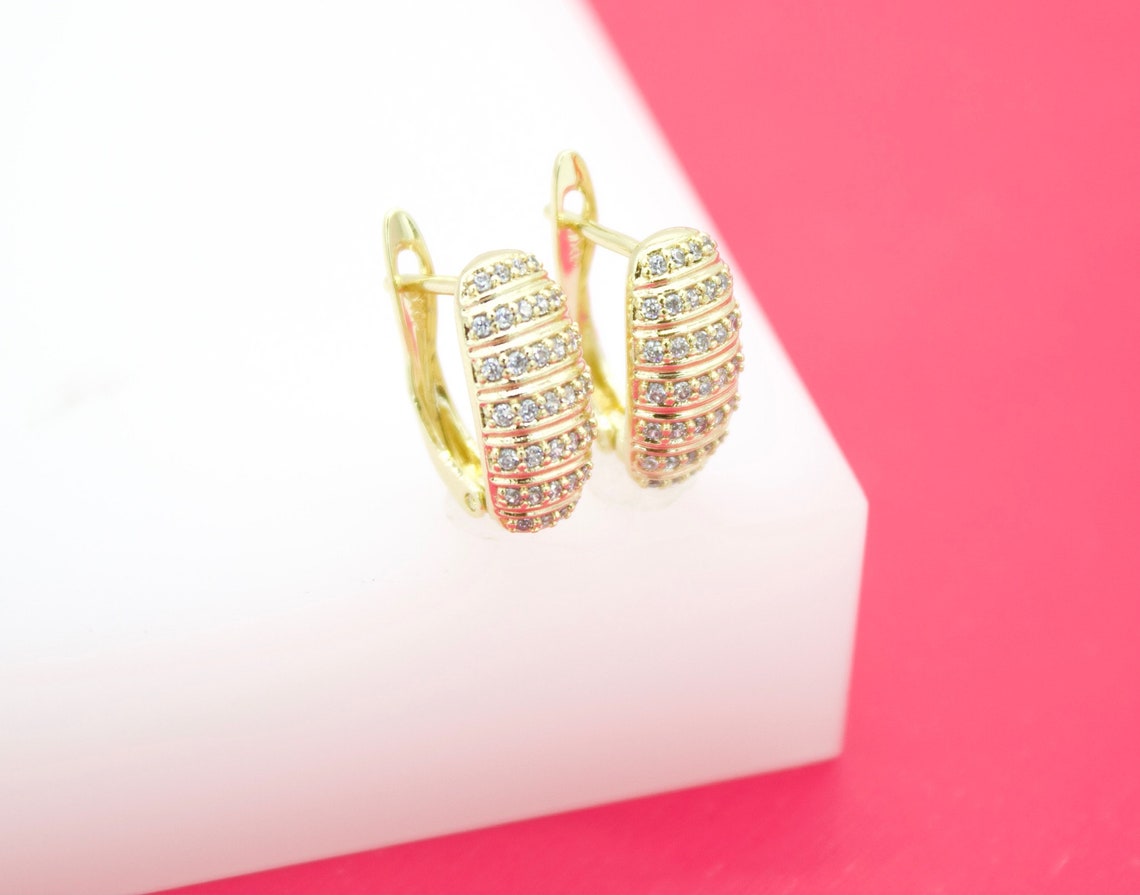 A pair of gold huggie hoop earrings featuring rows of sparkling cubic zirconia stones rests on a white display block against a pink background.