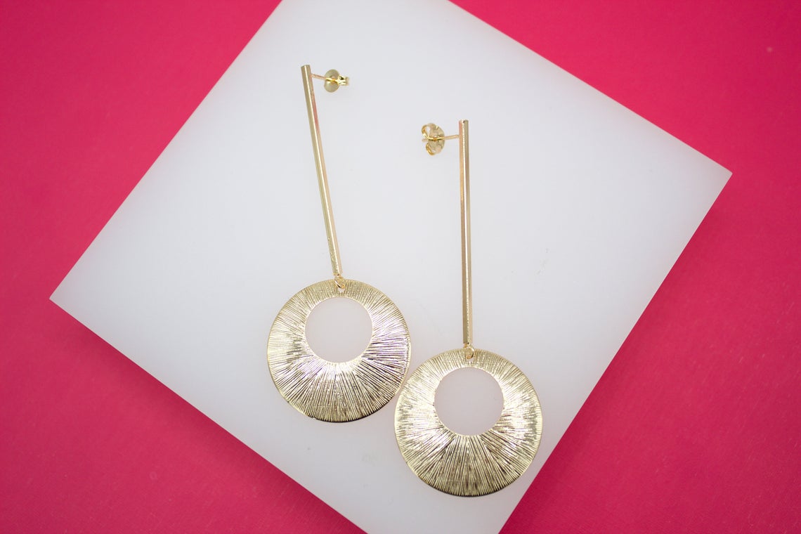 A pair of elegant long gold earrings featuring textured circular pendants rests on a white display against a vibrant pink background.