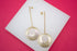 A pair of elegant long gold earrings featuring textured circular pendants rests on a white display against a vibrant pink background.