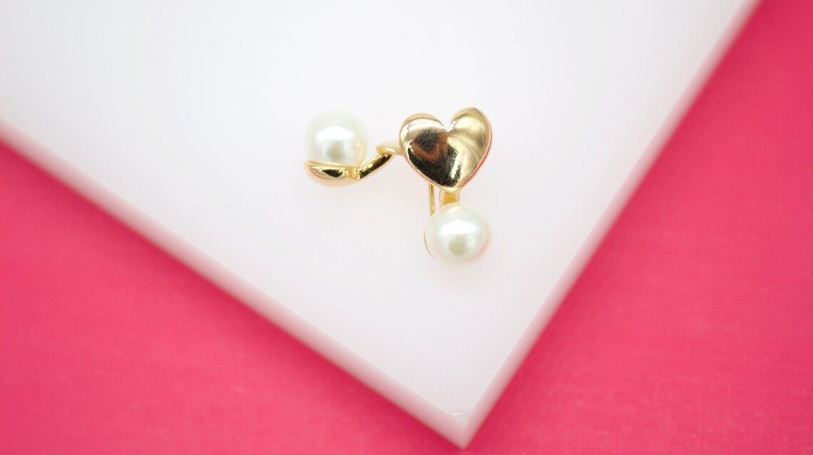 A gold heart-shaped stud earring with dangling white pearls rests on a white display against a vibrant pink background.