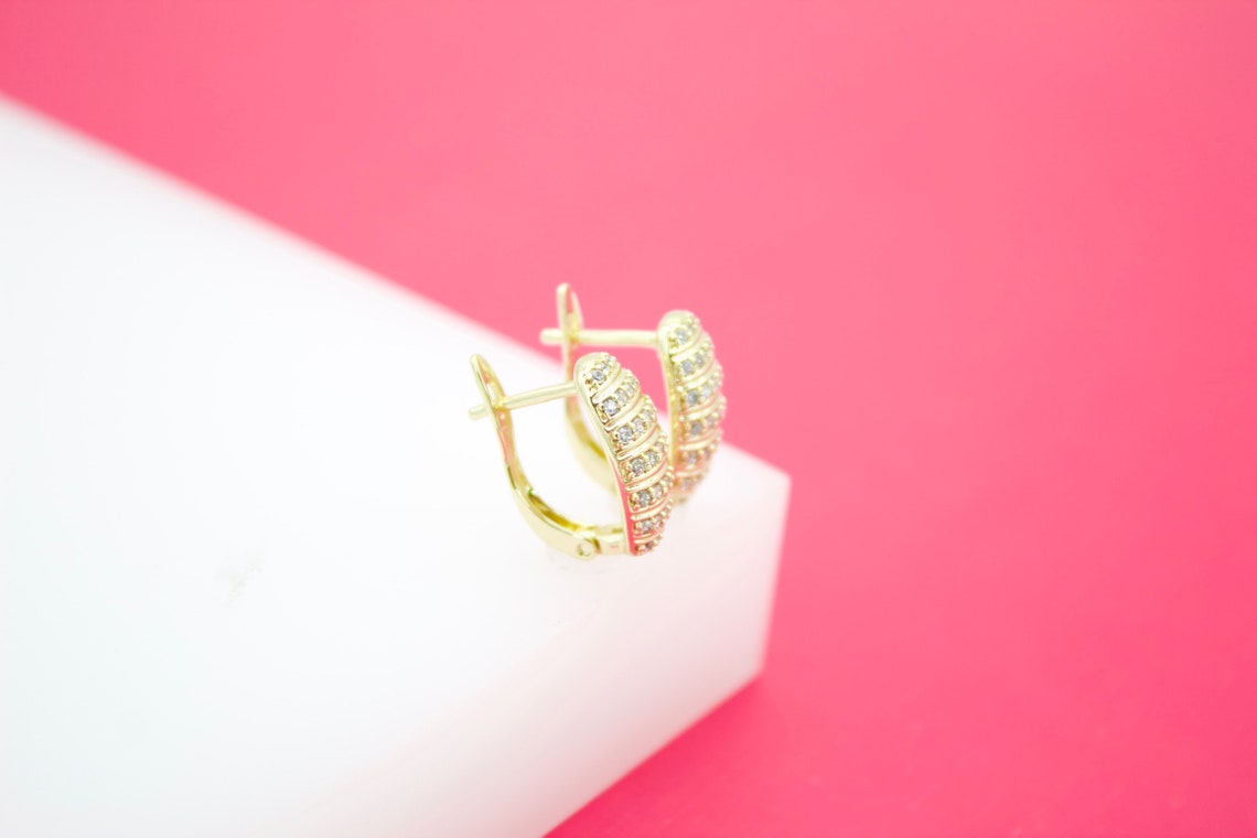 A pair of sparkling gold huggie hoop earrings encrusted with small crystals rests on a white block against a vibrant pink background.