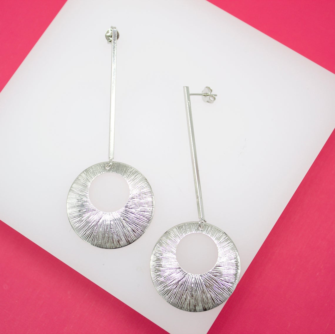 A pair of modern silver drop earrings with textured circular pendants rests on a white display surface against a vibrant pink background.