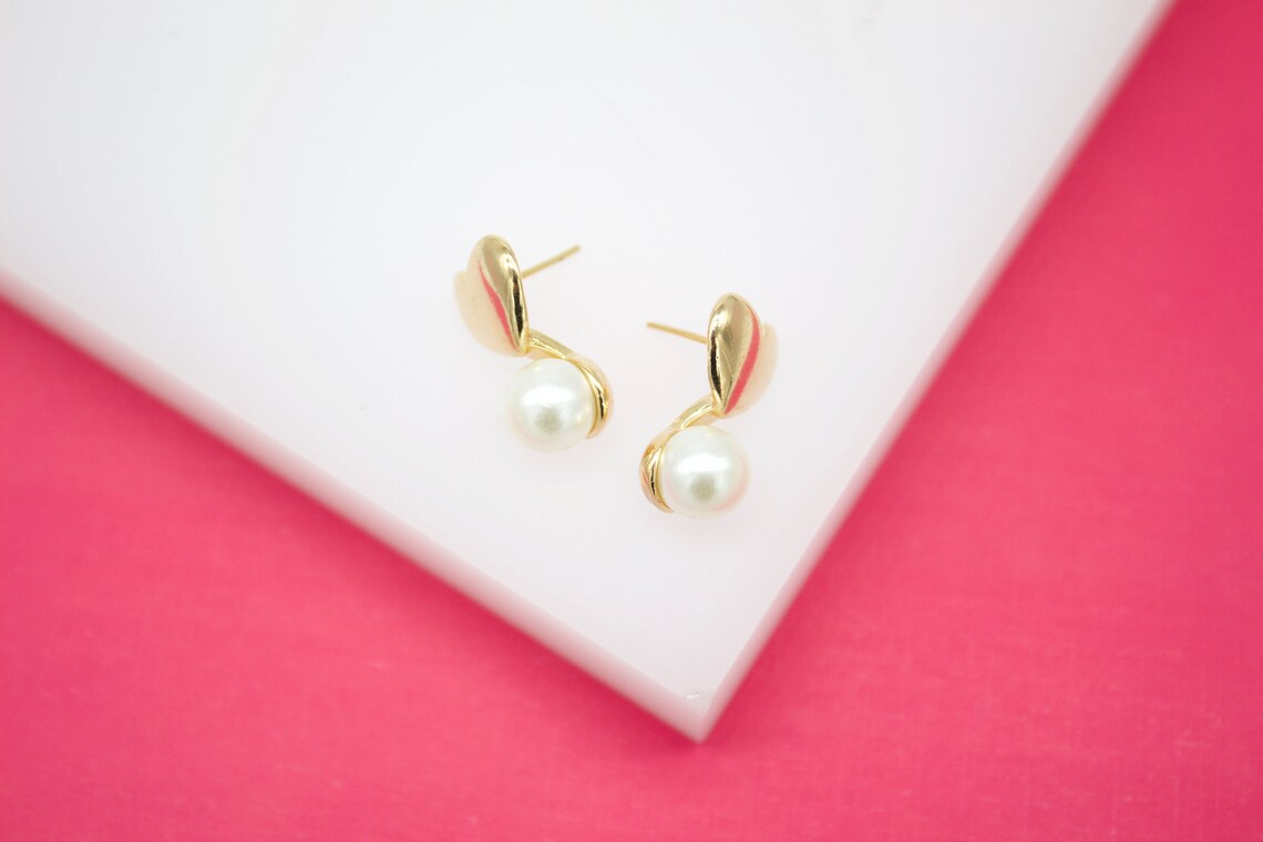 A pair of gold heart-shaped stud earrings featuring dangling white pearls rests on a white display surface against a vibrant pink background.