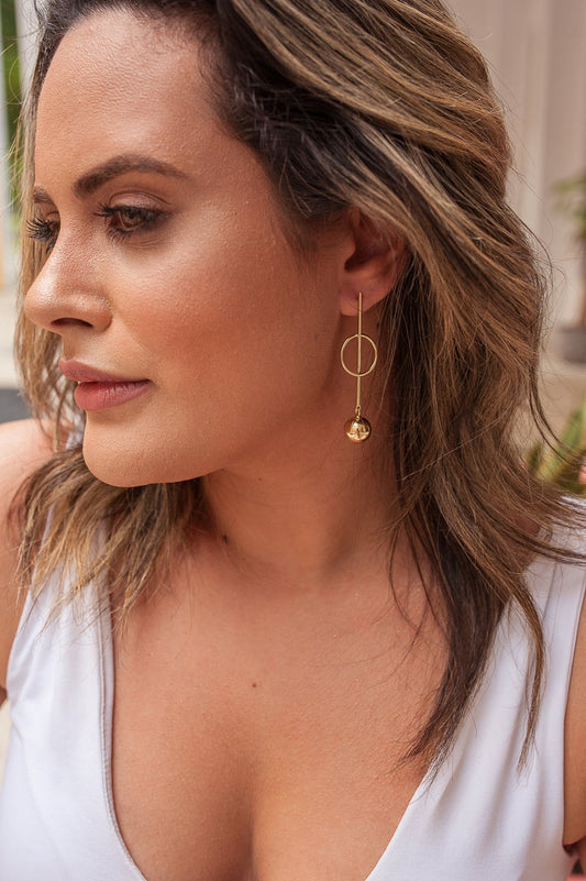 A woman wearing a modern geometric gold drop earring featuring a vertical bar, circle, and hanging sphere.