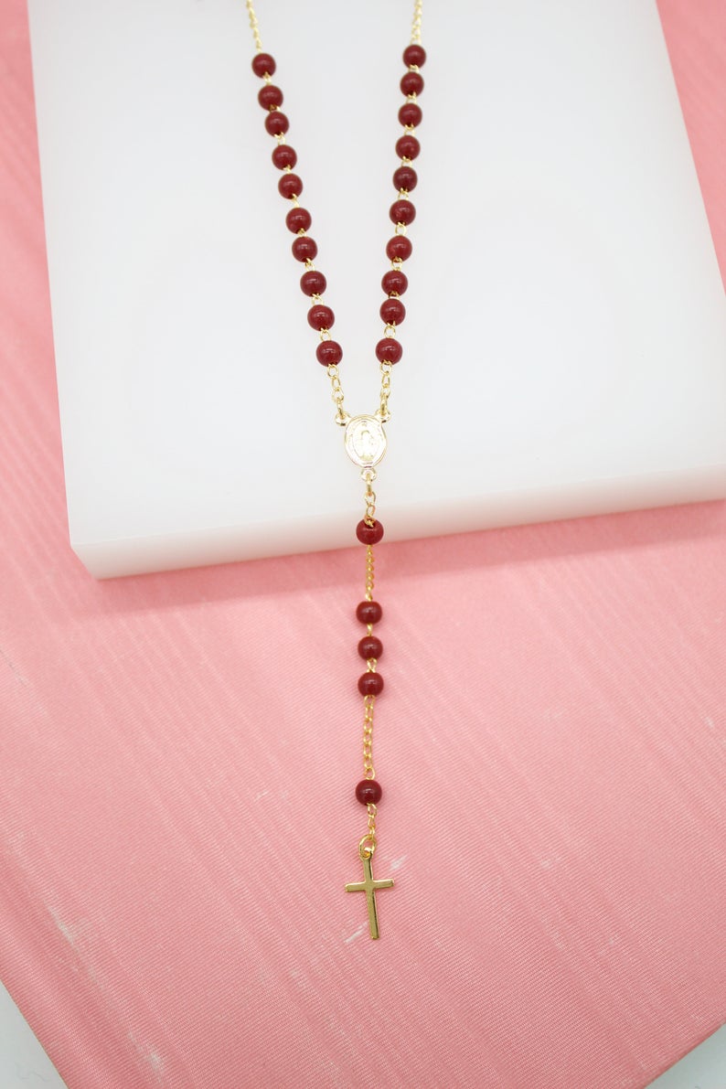 A delicate gold rosary necklace featuring deep red beads and a small crucifix rests on a white display against a pink background.
