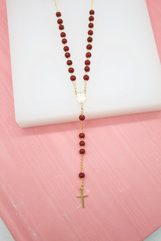 A delicate gold rosary necklace featuring deep red beads and a small crucifix rests on a white display against a pink background.