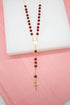 A delicate gold rosary necklace featuring deep red beads and a small crucifix rests on a white display against a pink background.