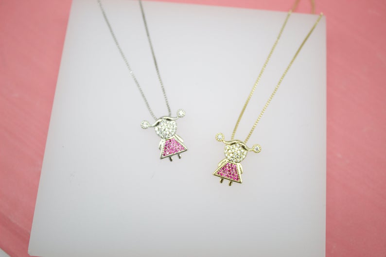 Two silver and gold little girl pendant necklaces with pavé-set crystals and pink dresses rest on a white display stand.