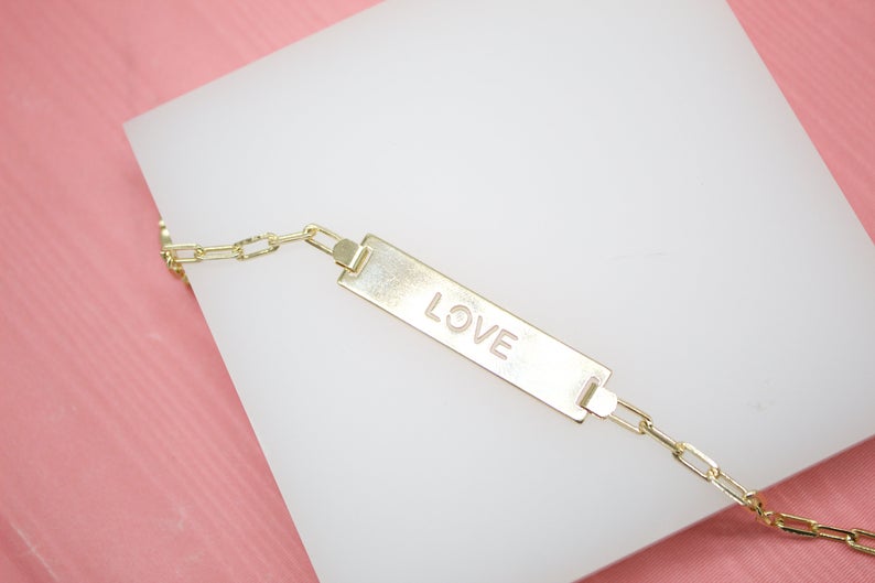A delicate gold ID bracelet featuring the engraved word "LOVE" rests on a white display block against a pink background.