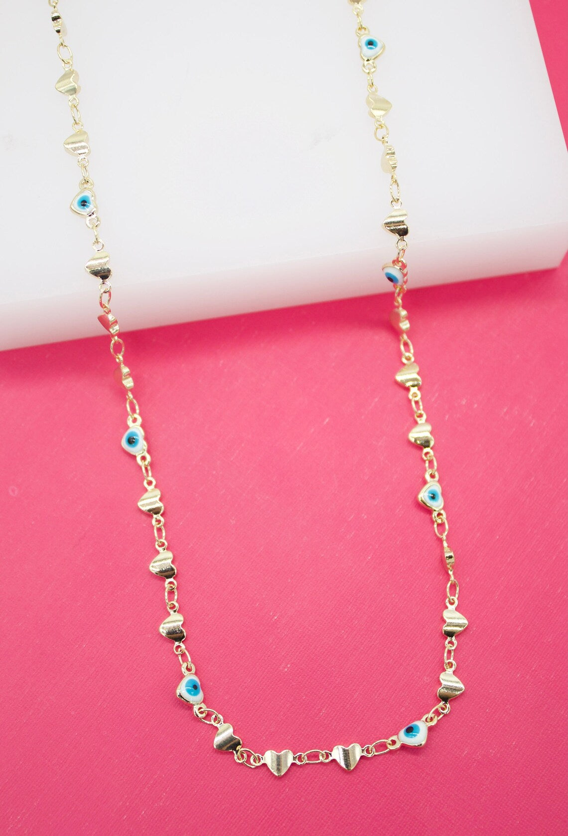 A delicate gold chain necklace drapes against a vibrant pink background, featuring alternating gold heart charms and tiny blue-and-white evil eye beads.