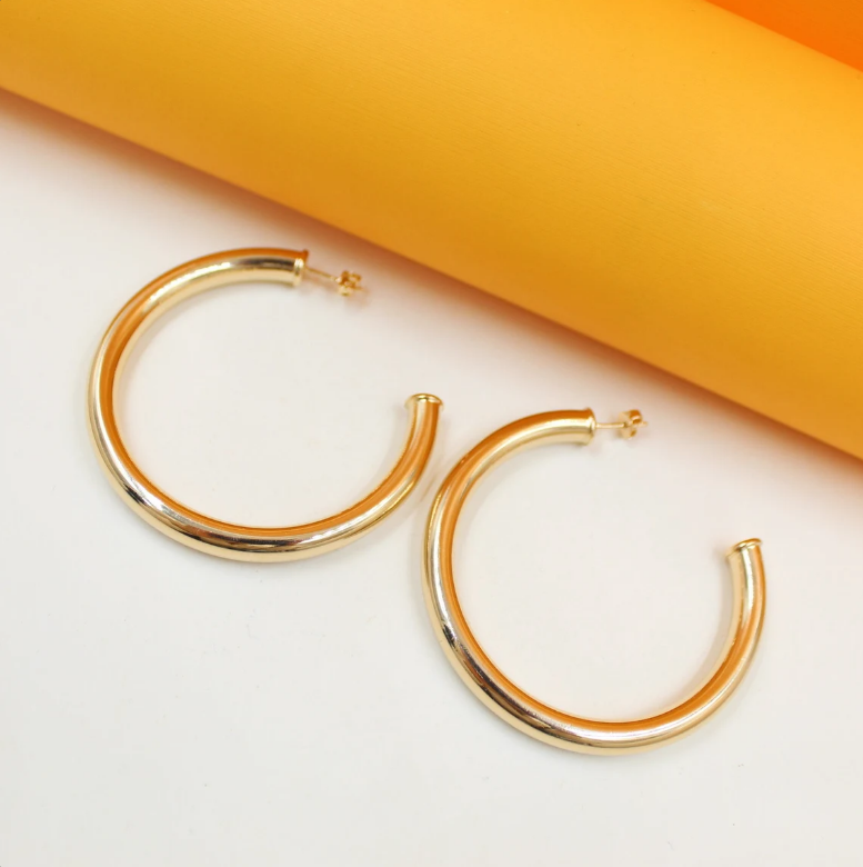 Two polished gold hoop earrings rest on a white surface against an orange backdrop.
