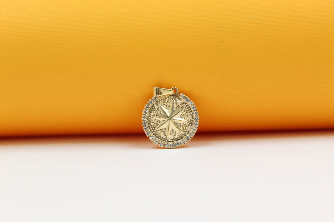 A gold pendant featuring a textured compass star design is encircled by sparkling cubic zirconia against a warm yellow and white backdrop.