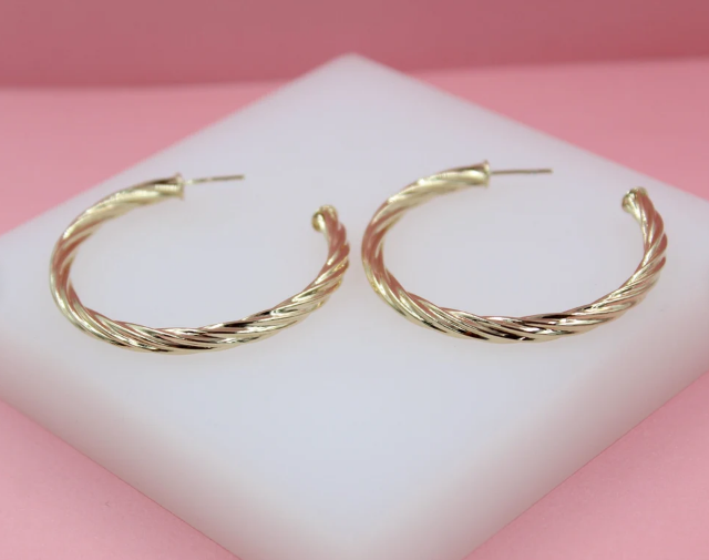 A pair of polished gold twist hoop earrings rests on a white display stand against a pink background.