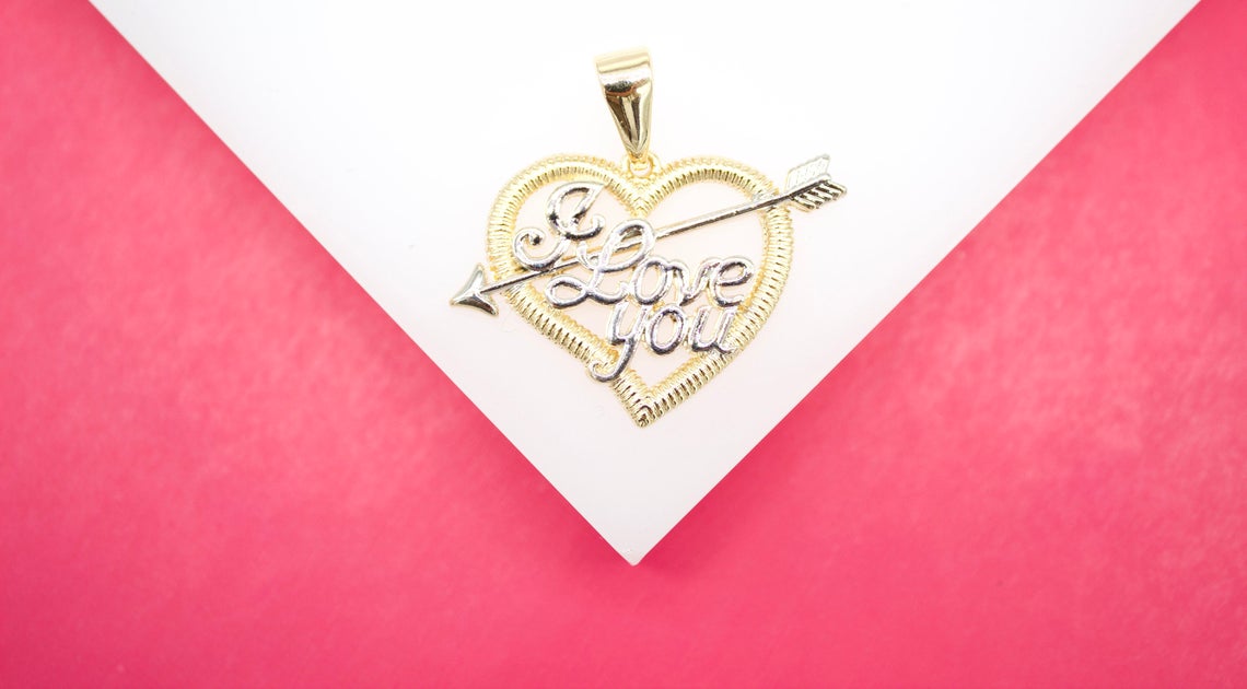A gold heart-shaped pendant featuring the inscription "I Love You" pierced by an arrow rests on a white display surface against a pink background.