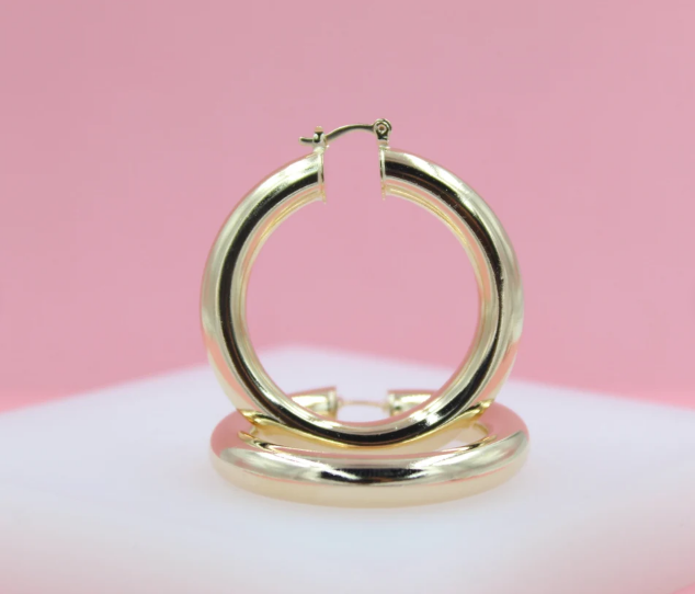 A pair of thick gold hoop earrings rests upright on a white platform against a soft pink background.