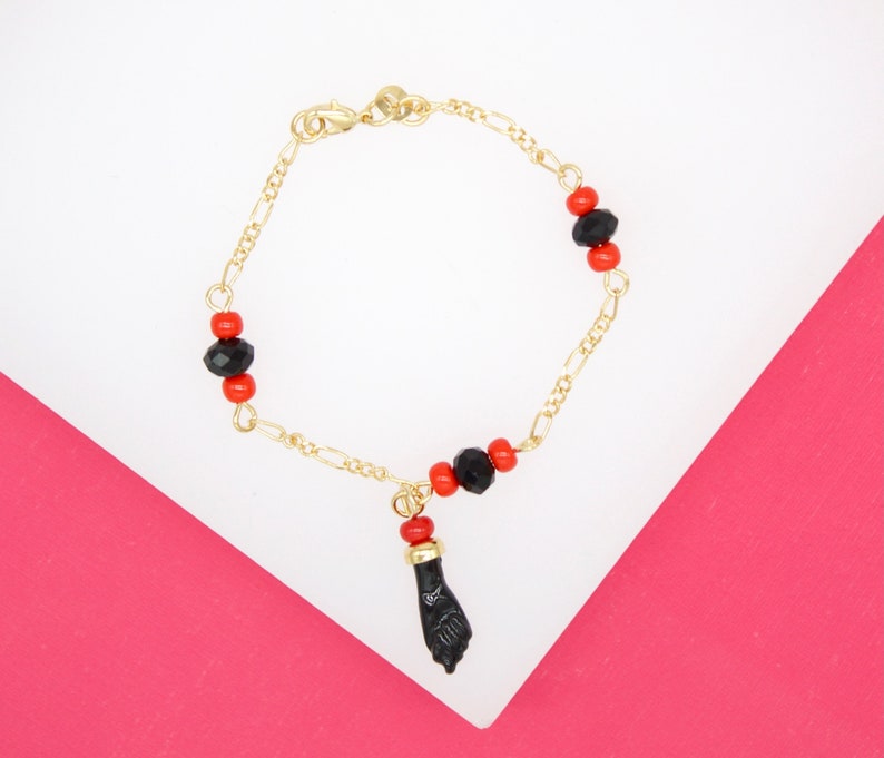 A delicate gold figaro chain bracelet features alternating red and black beads and hangs a black figa charm against a pink and white background.