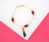 A delicate gold figaro chain bracelet features alternating red and black beads and hangs a black figa charm against a pink and white background.