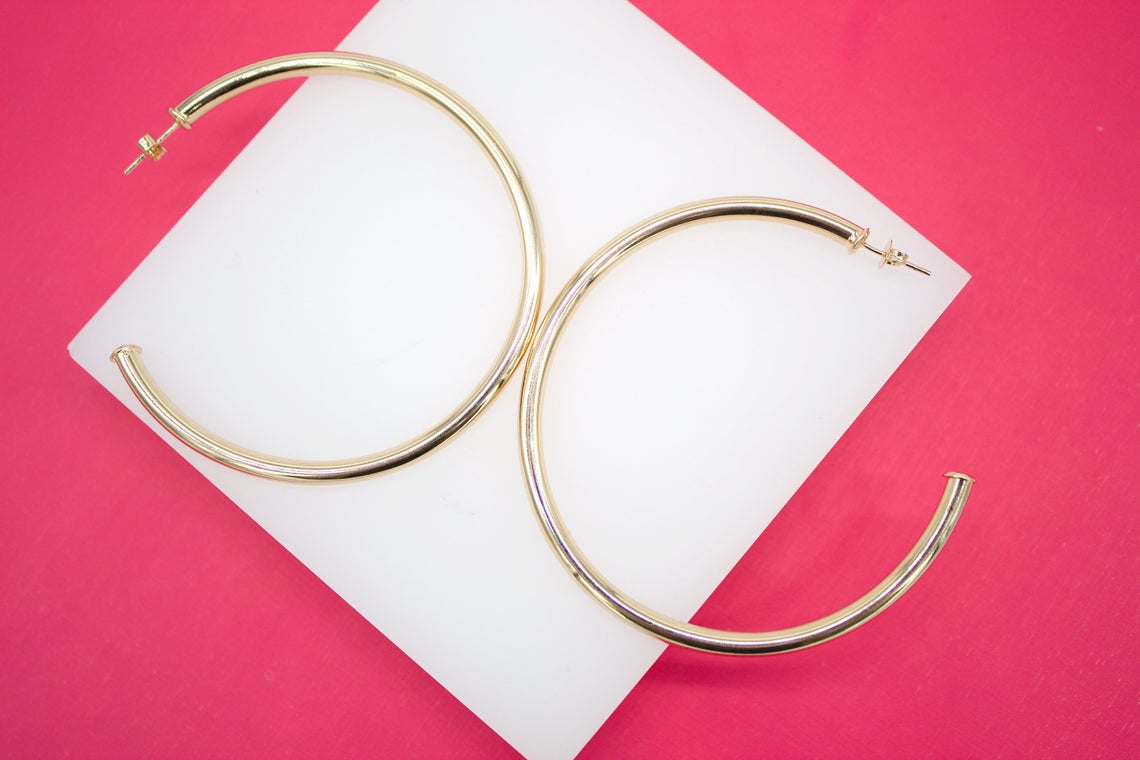 A pair of large, polished gold hoop earrings rests on a white display block against a vibrant pink background.