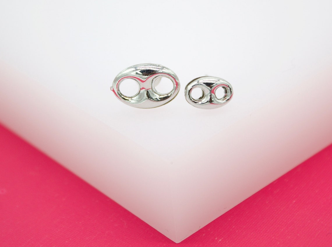 Two shiny silver puffed mariner link stud earrings rest side-by-side on a white display block against a vibrant pink background.