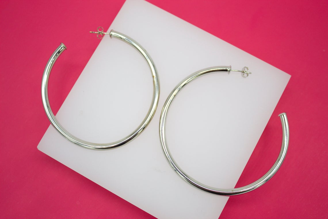 A pair of large silver hoop earrings rests on a white display block against a vibrant pink background.