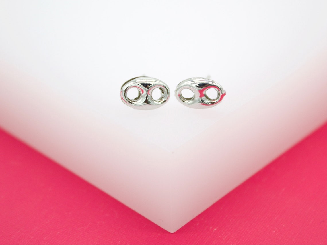 Two glossy silver mariner link stud earrings rest side-by-side on a white display surface against a vibrant pink background.