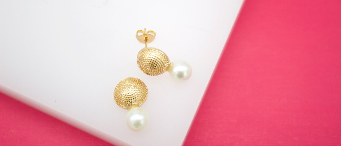 A pair of gold textured stud earrings with hanging white pearls rests on a white display against a pink background.