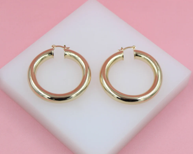 A pair of thick gold hoop earrings rests on a white display block against a pink background.
