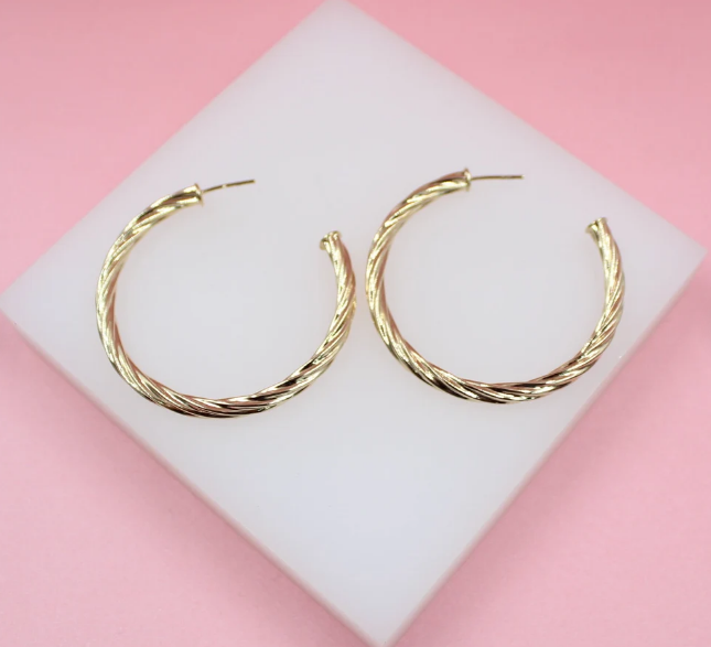 A pair of shiny gold twisted hoop earrings rests on a white block against a soft pink background.