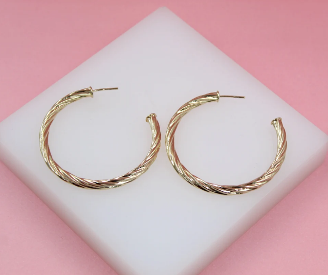 A pair of elegant gold twisted hoop earrings rests on a white display stand against a vibrant pink background.