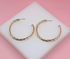 A pair of elegant gold twisted hoop earrings rests on a white display stand against a vibrant pink background.