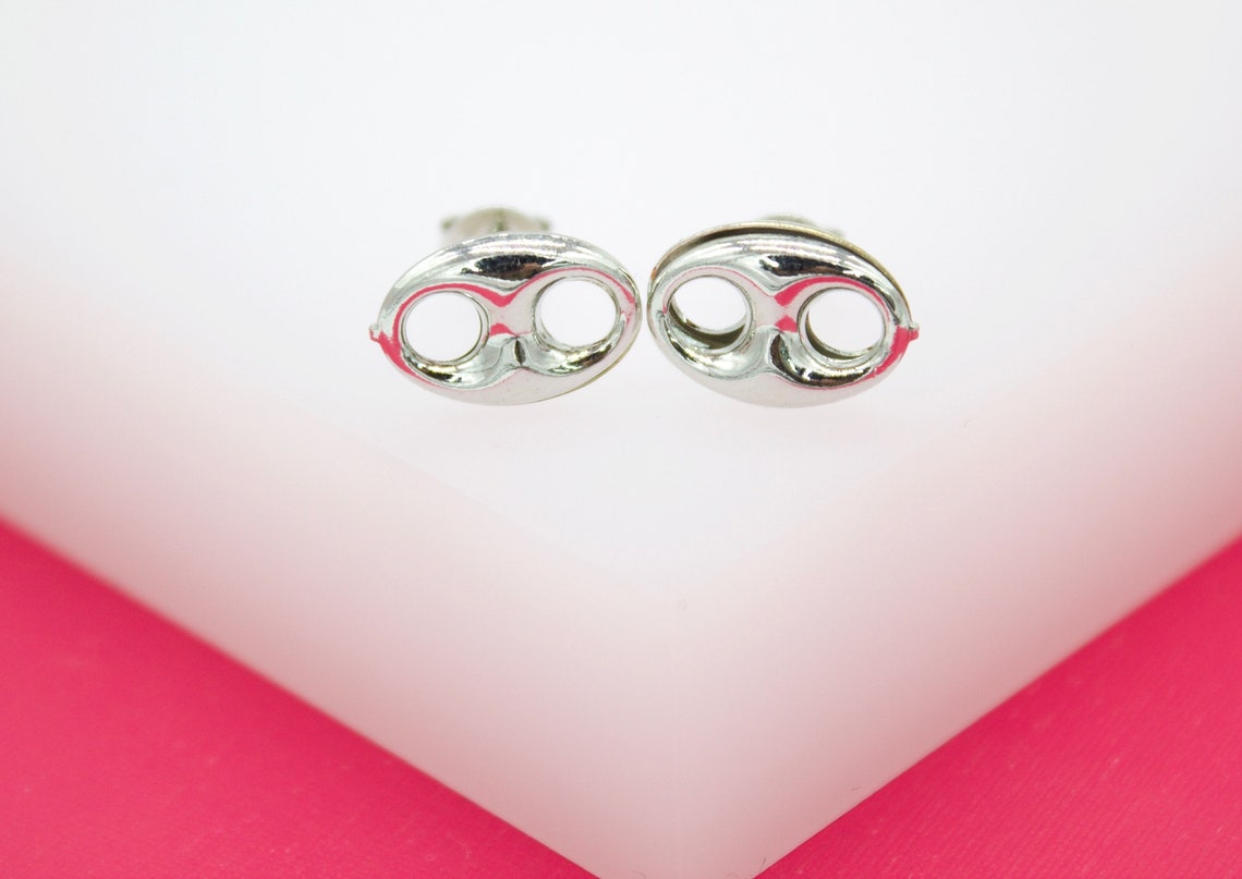 A pair of polished silver puff mariner anchor link stud earrings rests on a white display against a pink background.