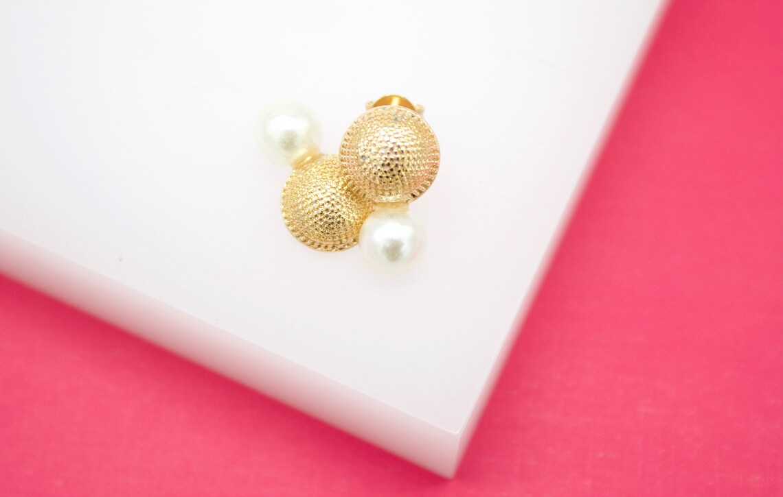 A pair of gold textured ball earrings with pearl accents rests on a white display surface against a pink background.