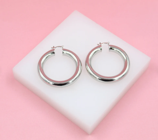 A pair of thick silver hoop earrings rests on a white display block against a soft pink background.