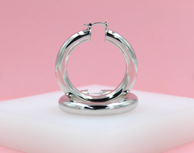 A pair of thick, polished silver hoop earrings rests on a white display block against a soft pink background.