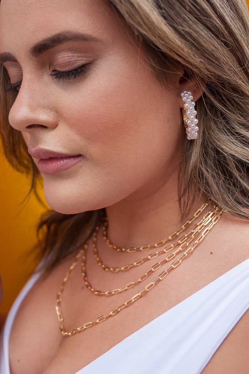 A blonde woman wearing chunky pearl hoop earrings and layered gold chain necklaces looks down thoughtfully.