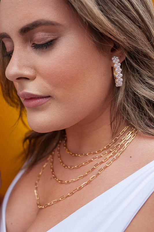A blonde woman wearing chunky pearl hoop earrings and layered gold chain necklaces looks down thoughtfully.