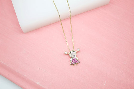 A delicate gold chain holds a sparkling little girl pendant featuring a pavé diamond head and a bright pink crystal dress against a soft pink backdrop.