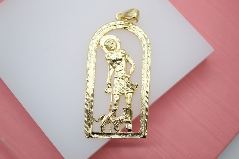 A gold San Lázaro pendant rests on a white display against a pink background, showcasing the intricate cutout figure of the saint accompanied by his dogs.