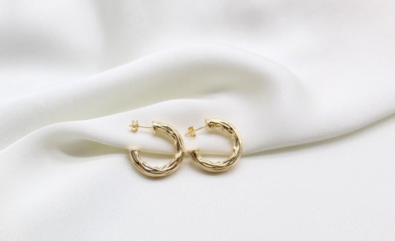 A pair of textured gold hoop earrings rests delicately on a soft white silk fabric background.