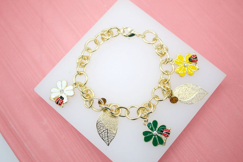 A gold charm bracelet featuring enamel flowers, leaves, and ladybug charms rests on a white display surface against a pink background.