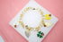 A gold charm bracelet featuring enamel flowers, leaves, and ladybug charms rests on a white display surface against a pink background.